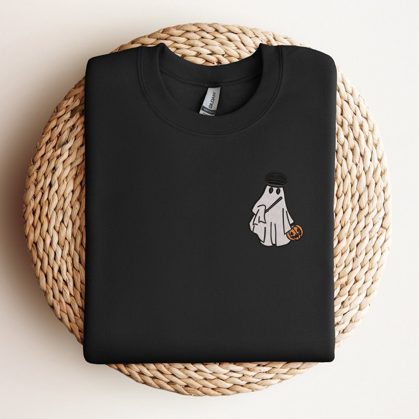Black sweatshirt with an embroidered design of a ghost wearing a police officer's hat and holding a pumpkin candy bucket, displayed on a woven mat.