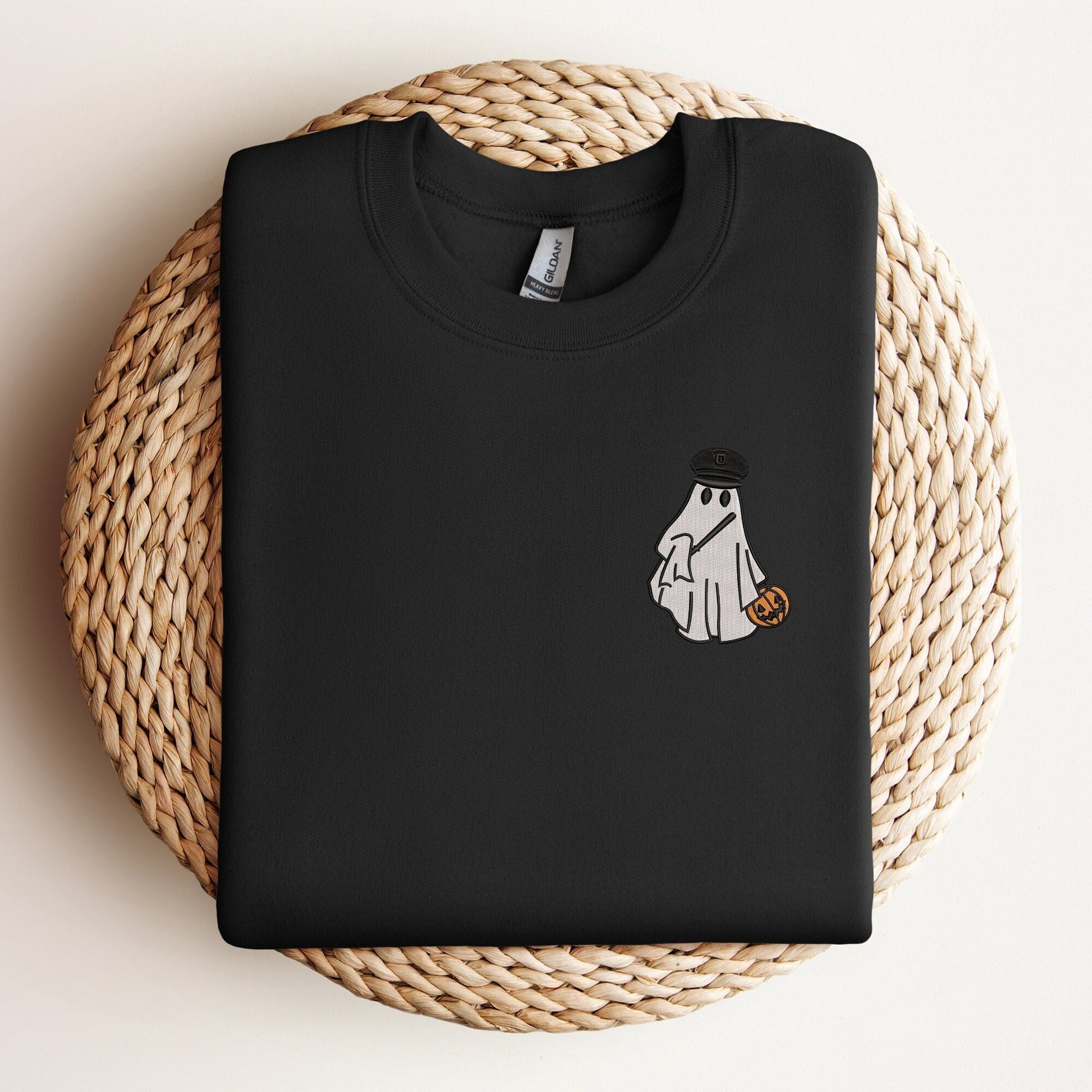 Black sweatshirt with an embroidered design of a ghost wearing a police officer's hat and holding a pumpkin candy bucket, displayed on a woven mat.